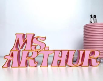 Könnte beinhalten: Rosa-goldenes Schreibtischschild mit der Aufschrift "Ms. Arthur". Die Buchstaben sind rosa mit goldenen Rändern. Im Hintergrund ist ein rosa Stifthalter mit Stiften zu sehen.