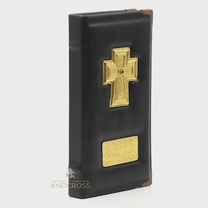 May include: Black leather-bound book with gold-colored cross and decorative accents. The cross features a small red gem in the center. The book has gold corner protectors and a gold rectangular plate with text.