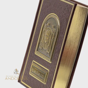May include: A brown leather-bound book with gold-gilded edges. The cover features a bronze-colored relief of a religious figure within an arched frame, and a rectangular plaque with text. The book has a textured pattern and gold trim.