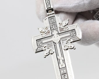 May include: A silver cross pendant with intricate details and engraved text in Cyrillic script. The cross features a central design with a smaller cross and a decorative border.