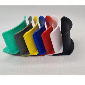 Könnte beinhalten: Ein Set aus sieben gebogenen, C-förmigen Kunststoffhaltern, die in einer Reihe angeordnet sind. Die Halter sind in den Farben Grün, Grau, Gelb, Blau, Rot, Weiß und Schwarz gehalten. Jedes Teil hat eine flache Basis und eine vertikale Wand.