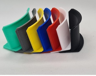 May include: A set of seven curved, C-shaped plastic holders arranged in a row. The holders are coloured green, grey, yellow, blue, red, white, and black. Each piece features a flat base and a vertical, semi-circular wall designed for support.