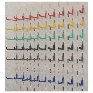 May include: A white pegboard panel featuring rows of multi-coloured J-shaped hooks. The hooks are arranged in horizontal lines, displaying a variety of colours including red, yellow, teal, black, grey, white, and blue, all attached to the board.