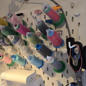 Può includere: Un pannello forato bianco come organizzatore a parete che sostiene diverse bobine di filo da cucito di vari colori, ciascuna racchiusa in una rete protettiva bianca. Le bobine sono montate su pioli individuali.