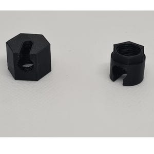 Könnte beinhalten: Zwei schwarze, sechseckige Bauteile mit einer zentralen Gewindeöffnung und einem vertikalen Schlitz an einer Seite. Die Objekte weisen eine durch 3D-Druck bedingte Schichtstruktur auf und verfügen über eine flache Oberseite sowie einen zylindrischen Sockel.