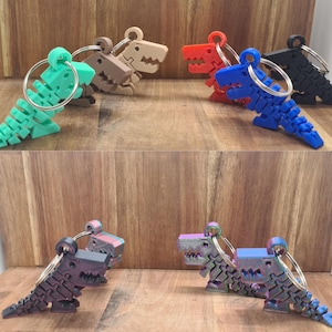 Op de afbeelding: Een collectie van gearticuleerde dinosaurus-sleutelhangers in diverse kleuren, waaronder mintgroen, beige, rood, blauw, zwart en iriserend paars. Elke sleutelhanger heeft een gesegmenteerd, flexibel lichaam bevestigd aan een ronde metalen ring.