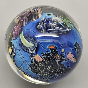 Può includere: Un fermacarte sferico in vetro trasparente con un design interno blu, nero e arancione. Il design presenta forme e motivi astratti, creando un aspetto artistico unico. Il fermacarte ha una superficie liscia e lucida.