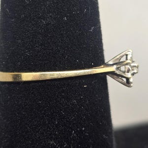 May include: A ring featuring a thin yellow gold band that transitions into a silver-toned metal setting. The setting holds a small, clear, faceted stone at the centre, secured by multiple prongs, viewed from a side profile perspective.