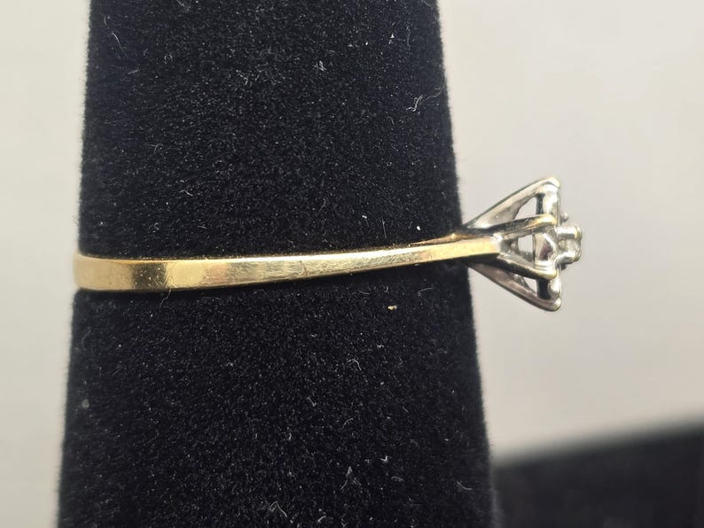 May include: A ring featuring a thin yellow gold band that transitions into a silver-toned metal setting. The setting holds a small, clear, faceted stone at the centre, secured by multiple prongs, viewed from a side profile perspective.