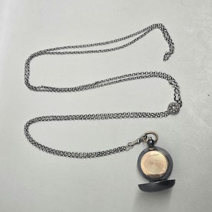 Puede incluir: Un collar plateado con un colgante redondo. El colgante tiene un interior dorado y un exterior gris oscuro con bisagras. La cadena del collar está hecha de pequeños círculos enlazados.