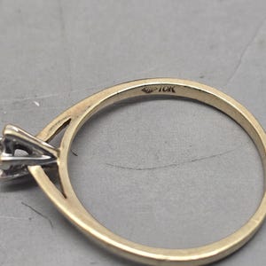 May include: A gold ring featuring a thin band and a silver-toned prong setting for a single stone. The interior of the band is clearly stamped with the text 10K, indicating the gold purity of the metal. The design includes a split shank detail.