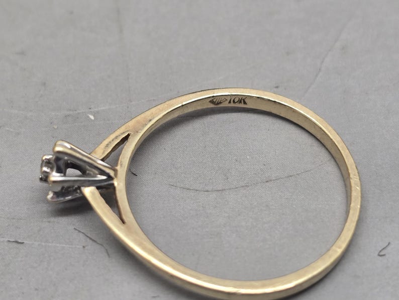 May include: A gold ring featuring a thin band and a silver-toned prong setting for a single stone. The interior of the band is clearly stamped with the text 10K, indicating the gold purity of the metal. The design includes a split shank detail.