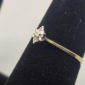 May include: A gold ring featuring a central setting with a small, clear, round-cut stone. The setting is shaped like a four-pointed star with metallic prongs holding the stone in place. The band is a simple, smooth, polished gold metal.