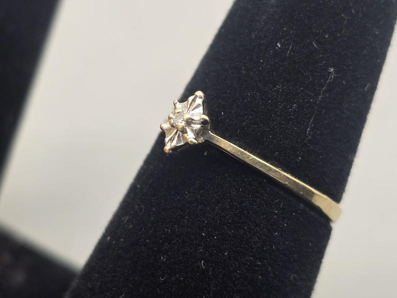 May include: A gold ring featuring a central setting with a small, clear, round-cut stone. The setting is shaped like a four-pointed star with metallic prongs holding the stone in place. The band is a simple, smooth, polished gold metal.