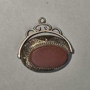 May include: A vintage gold-coloured pendant featuring an oval, reddish-brown stone. The pendant has an ornate, textured design around the stone and a decorative bail. The bail is a decorative loop at the top of the pendant.