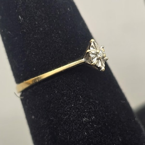 May include: A gold ring featuring a central diamond set within a star-shaped white gold or silver-toned setting. The band is smooth and yellow gold in colour, leading up to the elevated, multi-pointed metal mount that holds the small, clear stone.
