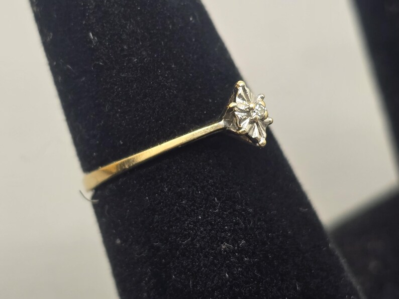 May include: A gold ring featuring a central diamond set within a star-shaped white gold or silver-toned setting. The band is smooth and yellow gold in colour, leading up to the elevated, multi-pointed metal mount that holds the small, clear stone.