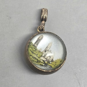 May include: A round pendant with a detailed miniature landscape scene. The pendant features a gold-toned metal frame and a clear, domed glass cover. The landscape includes green foliage, white structures, and a light blue sky.