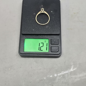 May include: A gold ring with a single stone setting resting on a black digital scale. The scale display shows a weight of 1.21 grams. The ring features a thin, polished gold band and a prong setting holding a small, clear, faceted gemstone.