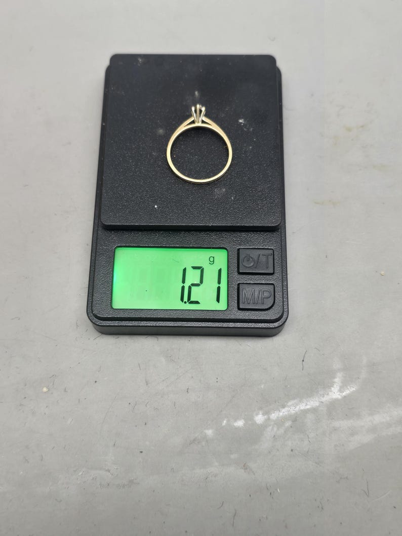 May include: A gold ring with a single stone setting resting on a black digital scale. The scale display shows a weight of 1.21 grams. The ring features a thin, polished gold band and a prong setting holding a small, clear, faceted gemstone.