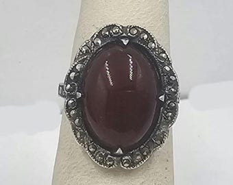 May include: A silver ring featuring a large oval red gemstone set in a detailed marcasite design.