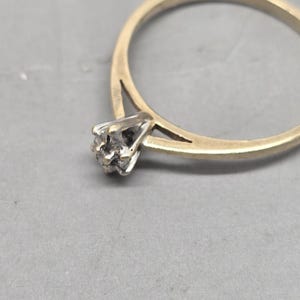 May include: A gold ring featuring a thin, polished band that splits as it reaches the setting. The centre stone is held in place by a silver-toned, multi-prong setting that elevates the small, clear gemstone above the band's surface.