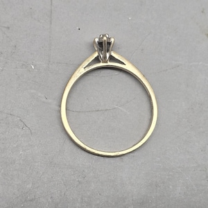May include: A gold ring featuring a thin, polished band that tapers towards a central setting. The setting is crafted from a lighter-toned metal and holds a single, small, dark-coloured stone secured by multiple prongs in a classic solitaire design.