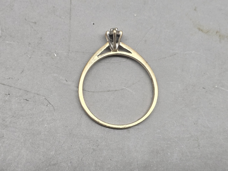 May include: A gold ring featuring a thin, polished band that tapers towards a central setting. The setting is crafted from a lighter-toned metal and holds a single, small, dark-coloured stone secured by multiple prongs in a classic solitaire design.