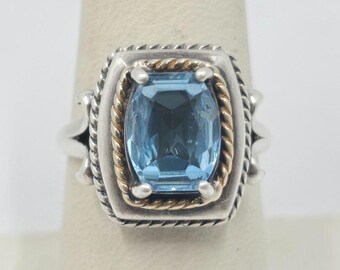 May include: A silver and gold ring featuring a large, light blue gemstone. The ring has a detailed, twisted rope design.