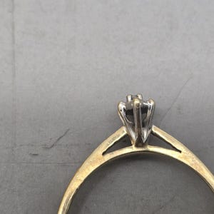 May include: A ring featuring a thin yellow gold band that transitions into a silver-toned metal setting. The setting holds a single small stone in a prong-style mount, creating a two-tone appearance with a smooth, polished finish on the metal.