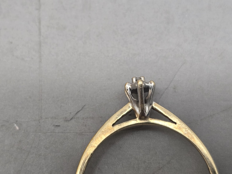 May include: A ring featuring a thin yellow gold band that transitions into a silver-toned metal setting. The setting holds a single small stone in a prong-style mount, creating a two-tone appearance with a smooth, polished finish on the metal.