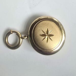 May include: A round gold-toned locket pendant featuring a brushed finish and a central engraved six-pointed star design. The locket is attached to a circular spring ring clasp, providing a secure mechanism for attachment to a chain or bracelet.