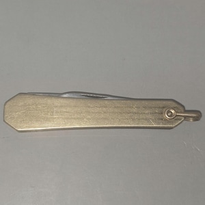 以下が含まれることがあります： 真鍮色のハンドルと銀色の刃が付いた、小型のヴィンテージポケットナイフ。ハンドルは長方形で、表面はテクスチャ加工されています。ハンドルの端には小さなループが付いています。