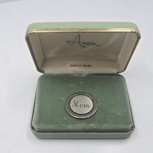 May include: A silver charm in a green velvet box with the word "Mom" engraved on it. The box is labeled "Anson Sterling Silver".