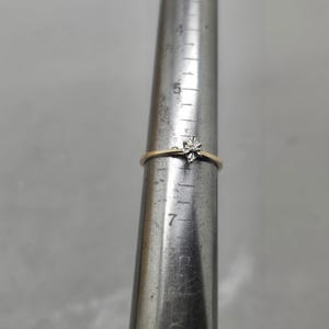 May include: A thin gold ring featuring a small, star-shaped silver-tone setting with a single clear stone. The ring is placed on a metal ring mandrel, indicating a size of approximately M on the standard British ring sizing scale.