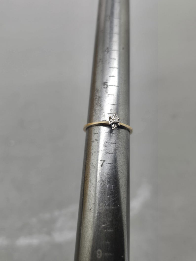 May include: A thin gold ring featuring a small, star-shaped silver-tone setting with a single clear stone. The ring is placed on a metal ring mandrel, indicating a size of approximately M on the standard British ring sizing scale.