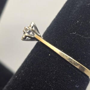 May include: A two-tone ring featuring a yellow gold band that transitions into a silver-toned setting. The setting holds a single, small, clear, faceted stone in a prong mount, showcasing a classic design with a polished metal finish.