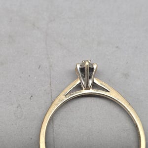 May include: A gold ring featuring a slender band and a six-prong setting that holds a single stone. The setting is crafted from a silver-toned metal, creating a contrast with the yellow gold band. The ring is shown from a side profile view.