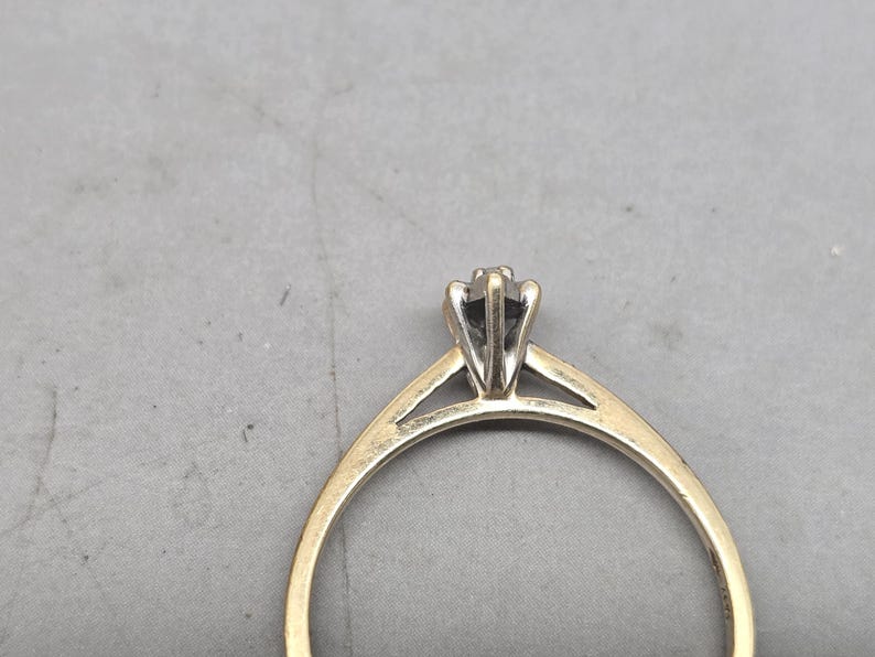 May include: A gold ring featuring a slender band and a six-prong setting that holds a single stone. The setting is crafted from a silver-toned metal, creating a contrast with the yellow gold band. The ring is shown from a side profile view.