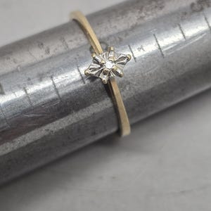 May include: A thin gold ring featuring a small, marquise-shaped setting with a central diamond accent. The ring is positioned on a metal ring mandrel, which displays measurement markings in centimetres, showing the band size clearly for reference.