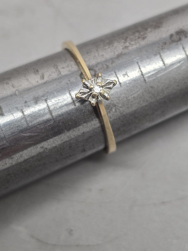 May include: A thin gold ring featuring a small, marquise-shaped setting with a central diamond accent. The ring is positioned on a metal ring mandrel, which displays measurement markings in centimetres, showing the band size clearly for reference.