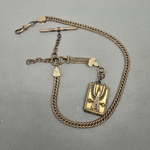 May include: Antique gold-toned pocket watch chain with a rectangular pendant featuring an Eiffel Tower design. The chain includes a T-bar, a clasp, and a decorative charm. The chain is made of a curb link style.