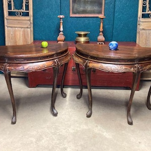 May include: Two antique, dark wood, half-moon tables with carved aprons and curved legs. Each table has a semi-circular top and is adorned with decorative carvings. A green apple and a blue and white ball sit on top of the tables.
