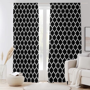 以下が含まれることがあります： 白と黒の模様のカーテン。カーテンは、黒い背景に白い幾何学模様が繰り返されています。窓の前に吊るされており、背景にはソファやその他の装飾が見えます。