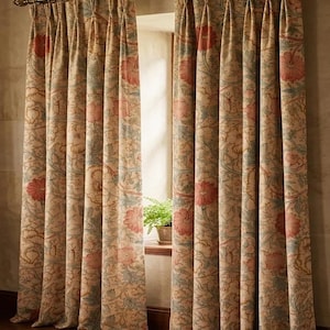Op de afbeelding: Beige gordijnen met bloemenpatroon en een goudkleurige stang. De gordijnen hebben een herhalend patroon van roze bloemen en groene bladeren. De gordijnen hangen voor een raam.