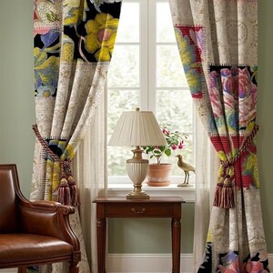 Op de afbeelding: Een paar kamerhoge gordijnen met een veelkleurig patchwork bloemenpatroon in tinten geel, rood, blauw en zwart. De gordijnen worden bijeengehouden door gevlochten koordhouders met kwastjes en hangen aan een metalen roede voor een raam.