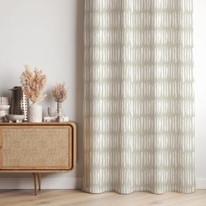 Puede incluir: Una cortina verde salvia con un patrón repetitivo de pinceladas blancas verticales. La cortina cuelga frente a una pared blanca. A la izquierda se ve un mueble de madera con objetos decorativos.