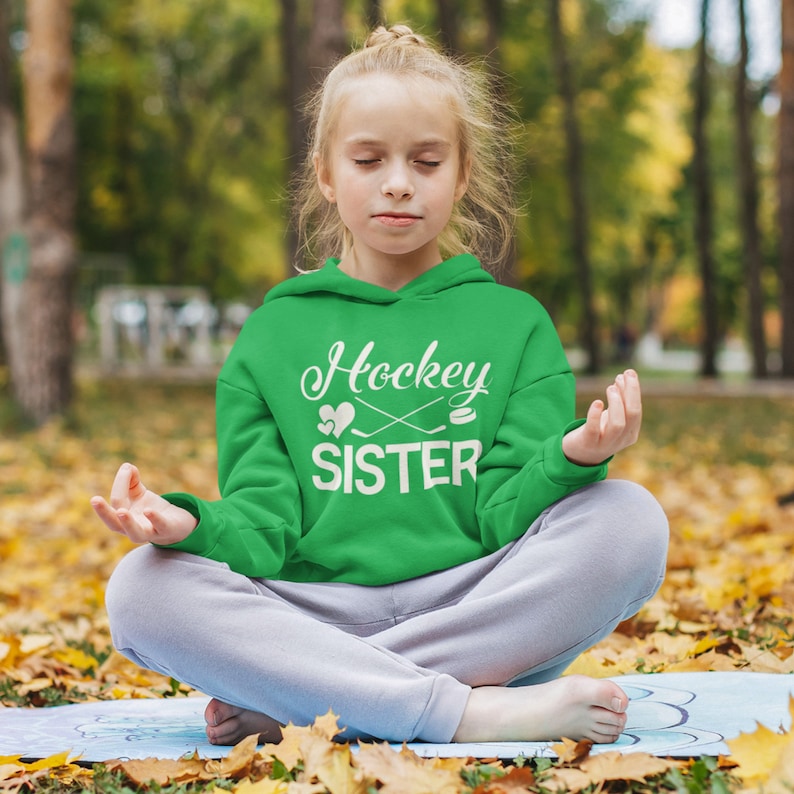 May include: A young person wearing a green hoodie with the text "Hockey Sister" and a hockey stick and puck graphic. The person is sitting cross-legged on a blue yoga mat with fall leaves around them.