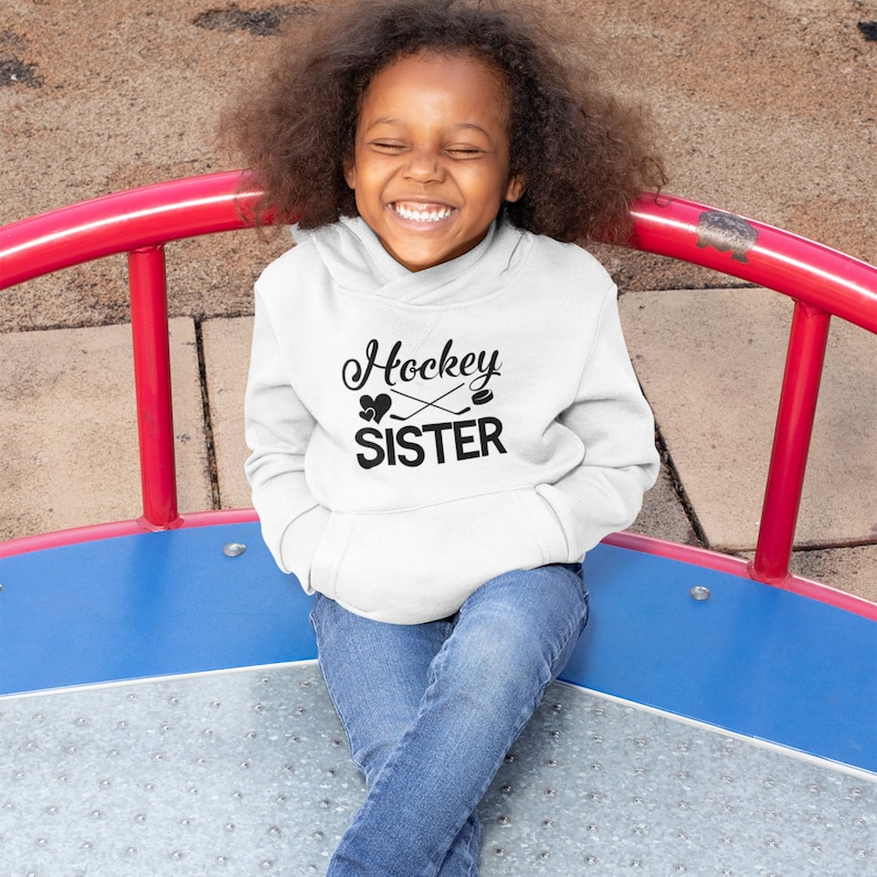 May include: A white hoodie with black text that says "Hockey Sister" with a hockey stick and puck graphic.