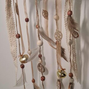 May include: A handcrafted dream catcher with a woven twig base. It features hanging ribbons, feathers, wooden beads, gold bells, and decorative wooden cutouts. The color palette includes natural browns, creams, and gold.
